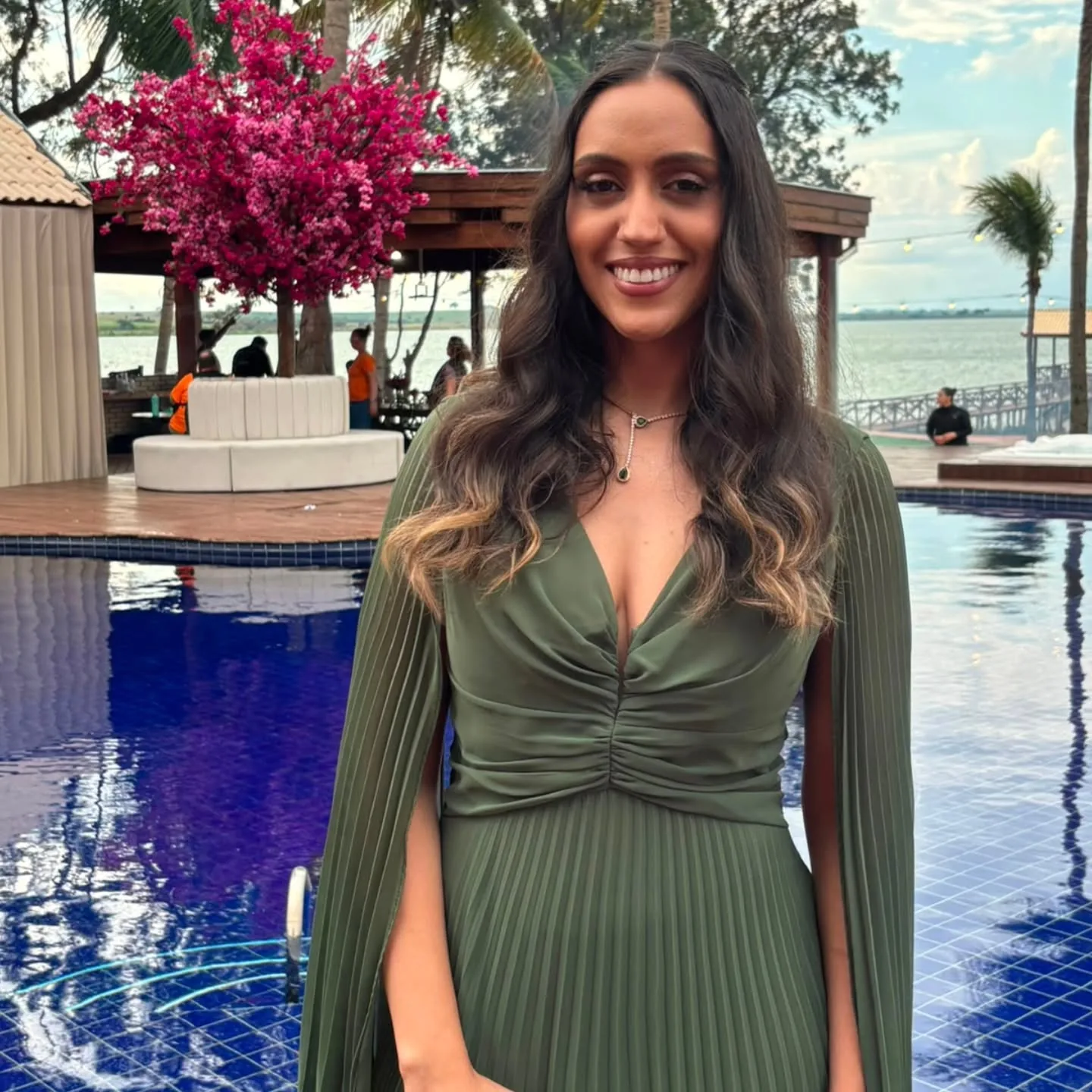 Thayane Alves de vestido longo verde, sorrindo, ao pôr do sol junto a uma piscina